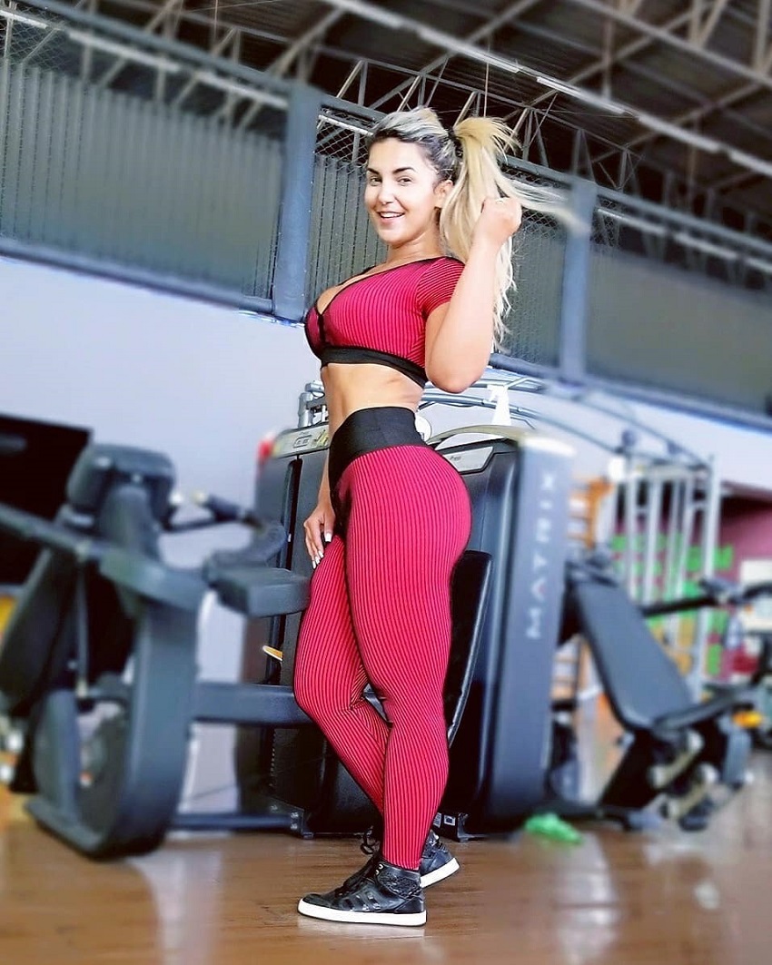 Daiane Lopes standing in a gym and smiling at the camera looking curvy and fit in her red sportswear