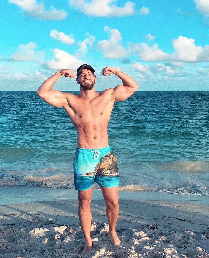 Darian Alvarez standing on the shore near a vast sea during a sunset, flexing his biceps for a photo