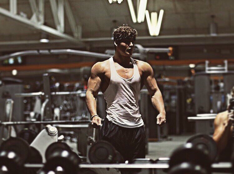 Justin Martilini standing in a gym by a dumbbell rack looking at himself in a mirror