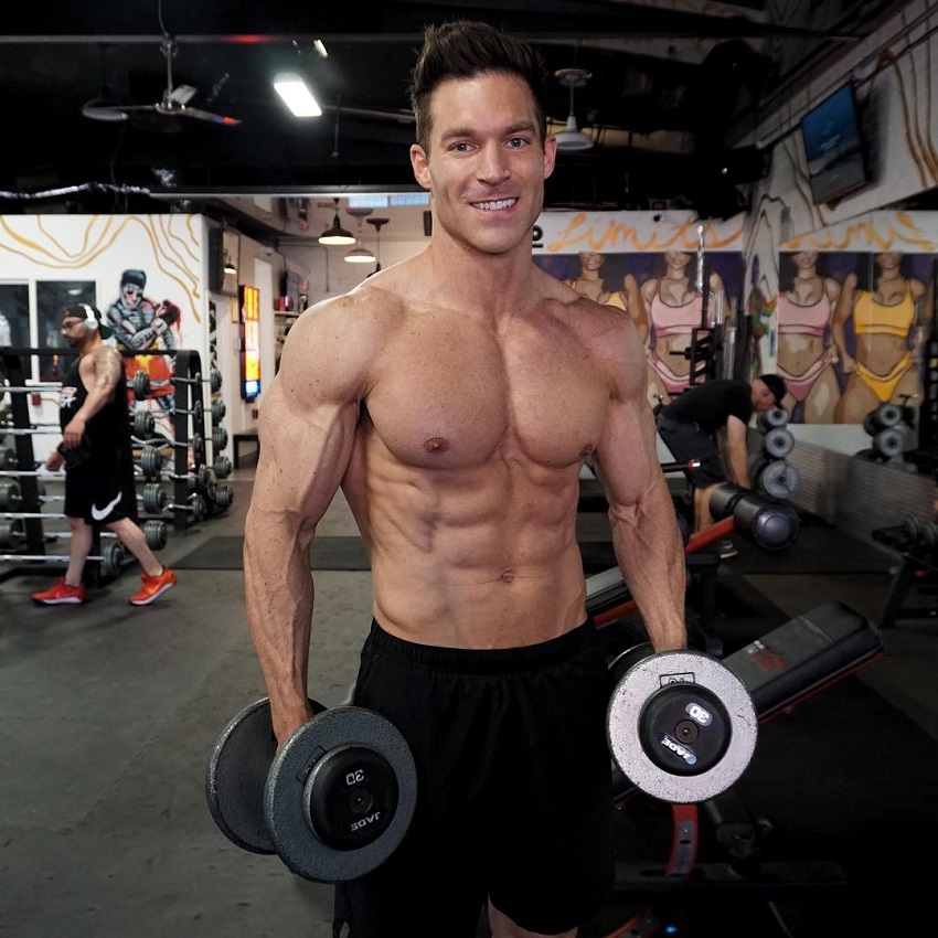 Kyle Clarke standing shirtless in the gym holding dumbbells in his hands