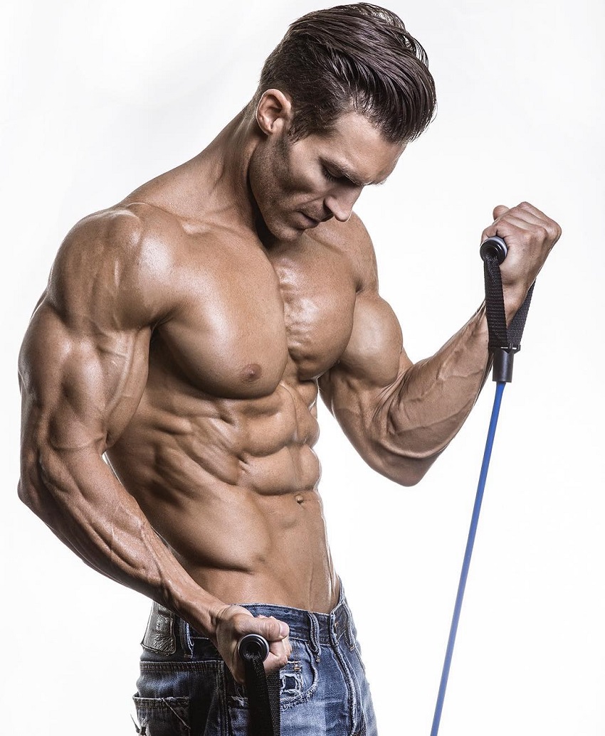 Kyle Clarke performing resistance band bicep exercise in a photo shoot, showing off his ripped and muscular body