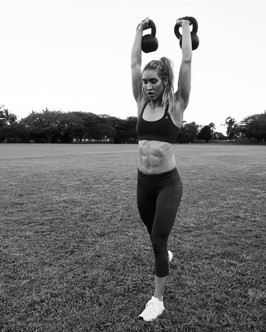 Cass Olholm doing overhead press with kettlebells on a grass field outdoors