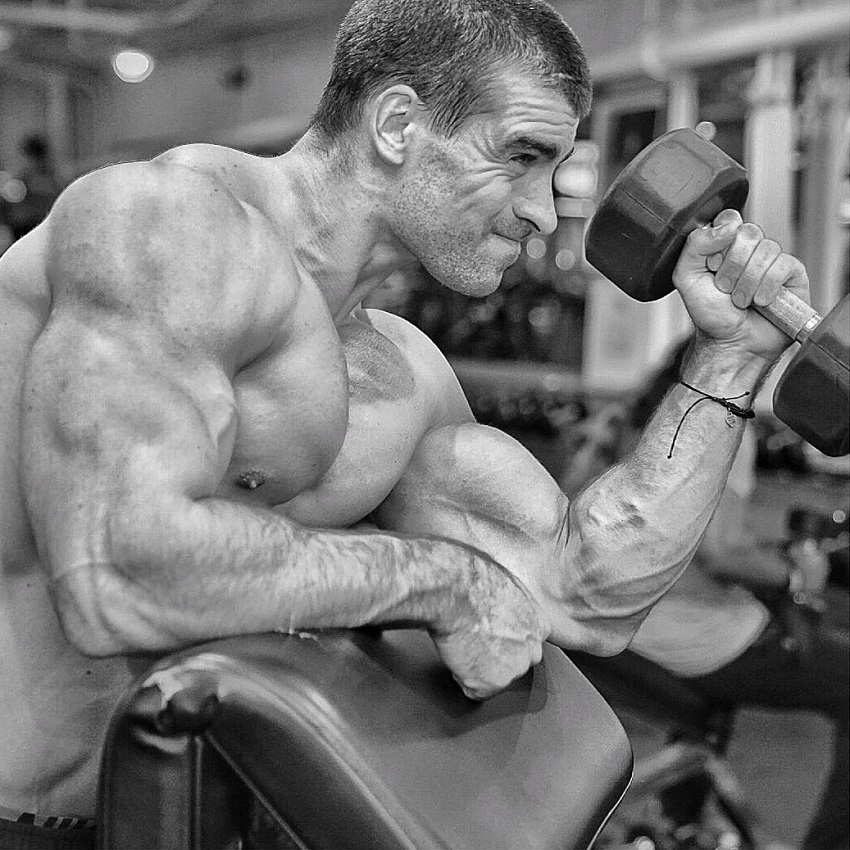 Thomas Canestraro doing heavy dumbbell biceps curls while being shirtless, looking strong and ripped