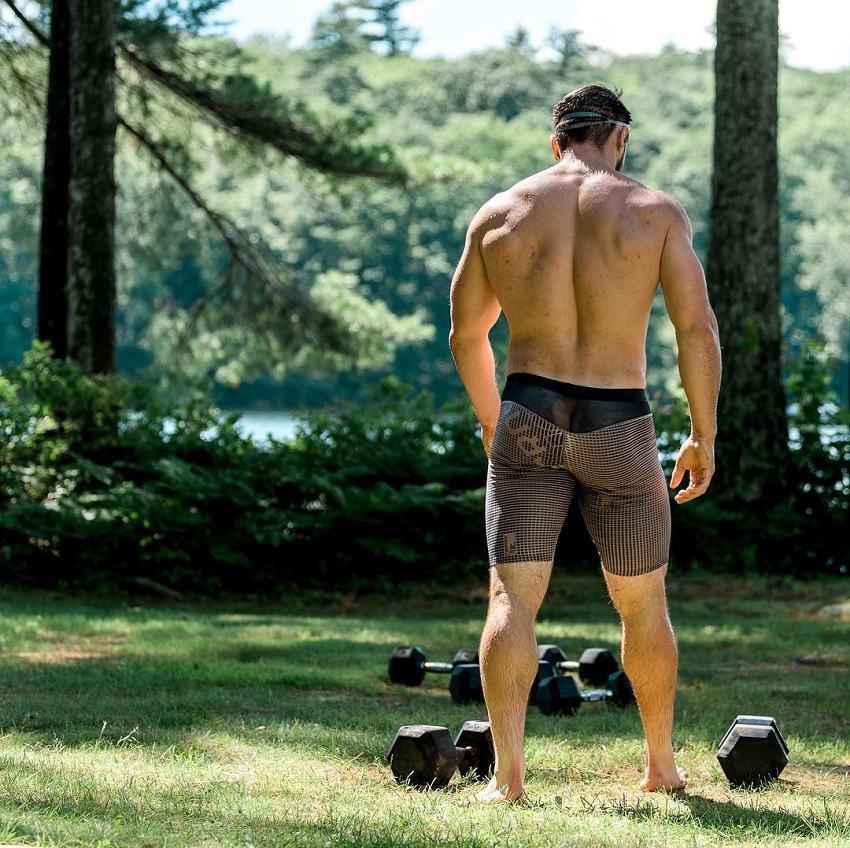 Travis Williams standing in the nature with trees and dumbbells on the grass, his back looking muscular and lean