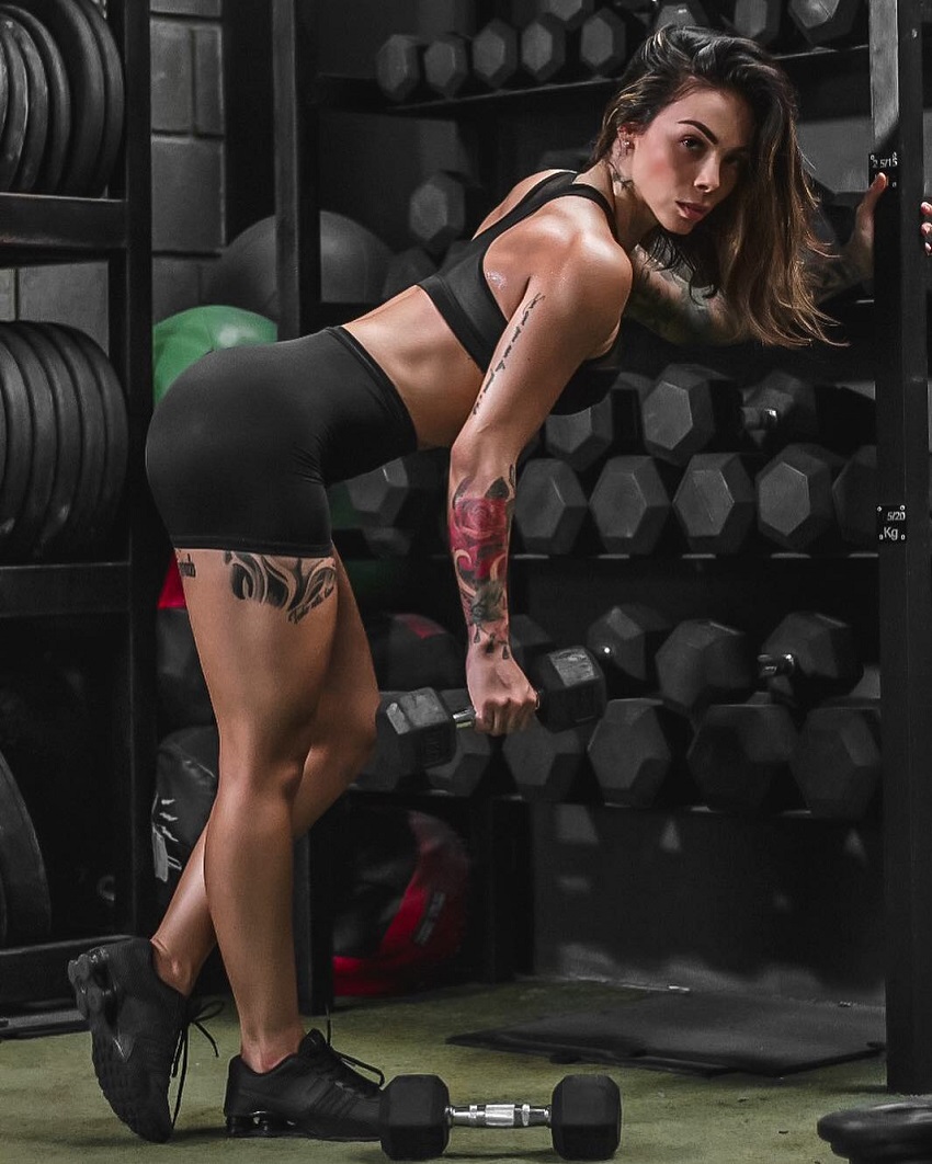 Angelica Fernandez posing by a dumbbell rack with a dumbbell in her hand