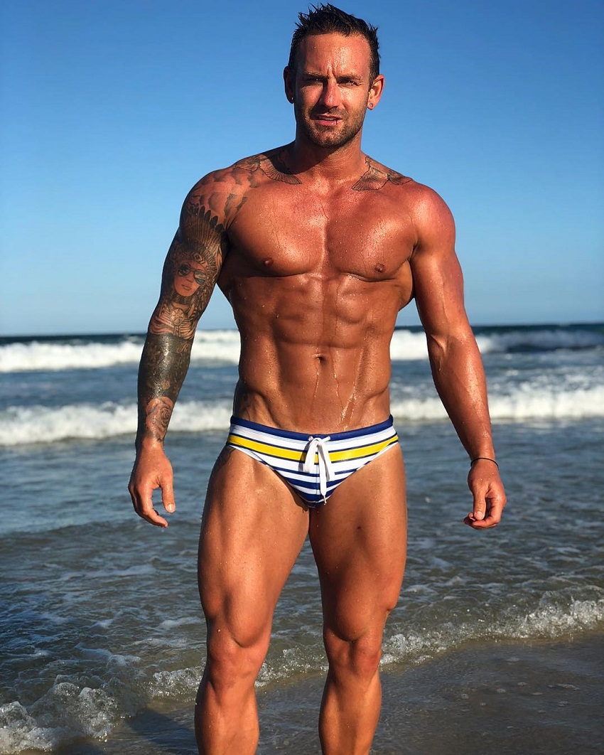 Joel Bushby posing shirtless for a photo on the beach, showing off his muscular upper body and legs