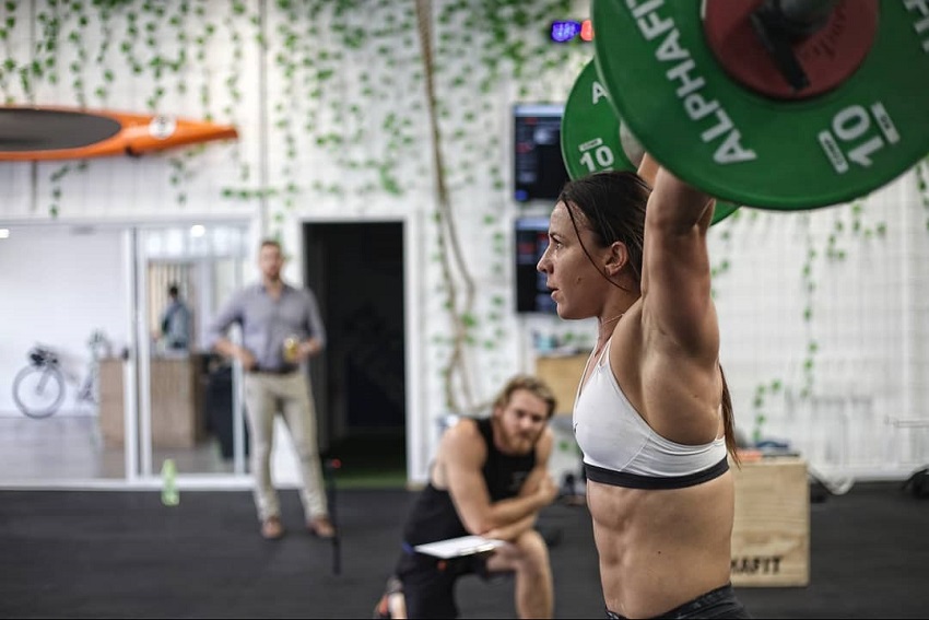 Kara Webb Saunders doing an overhead press with a barbell, looking fit and strong