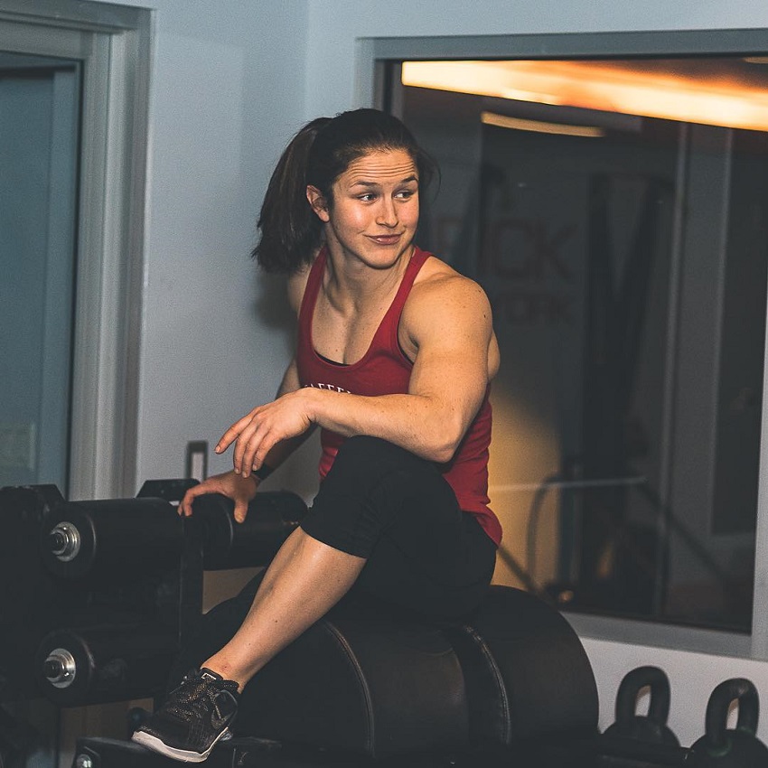 Kari Pearce sitting on a dumbbell rack looking fit and strong