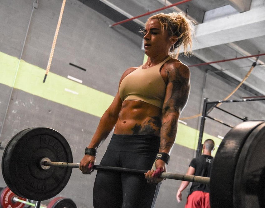 Lauren Herrera lifting a heavy barbell loaded with weights, her abs popping out