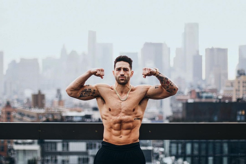 Noah Neiman doing a shirtless front double biceps flex for a photo with a huge city in the background