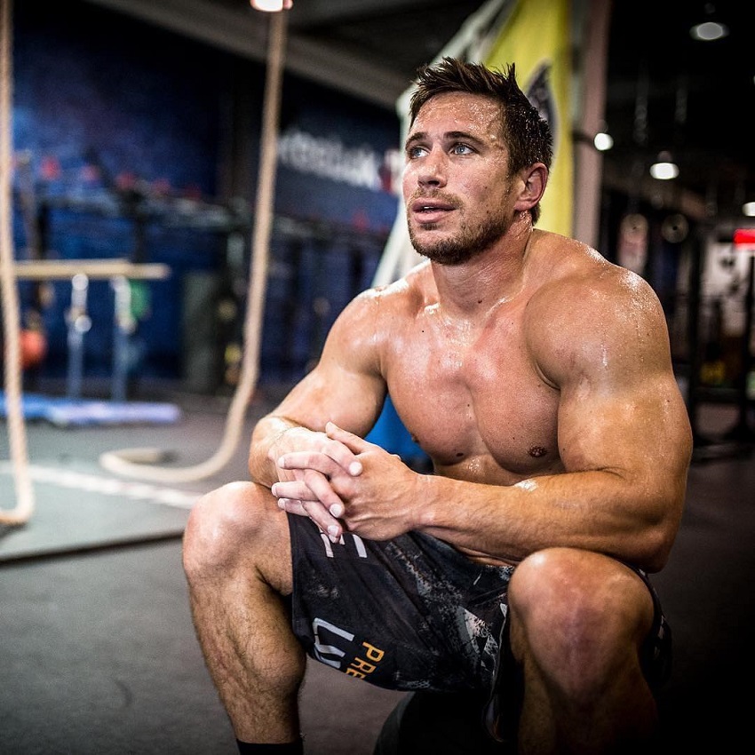 Dan Bailey sitting shirtless in a CrossFit training room, looking strong and muscular