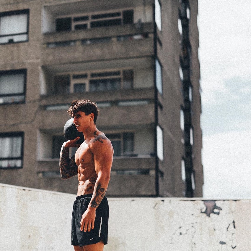 Adi Gillespie standing shirtless on top of a building with a black medicine ball in his hand