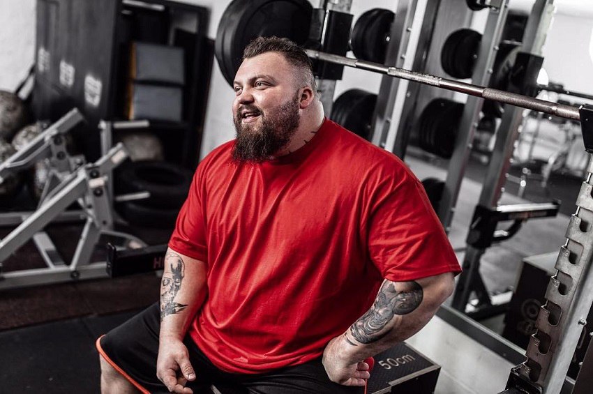 Eddie Hall sitting by the bench press in his red shirt looking big and strong