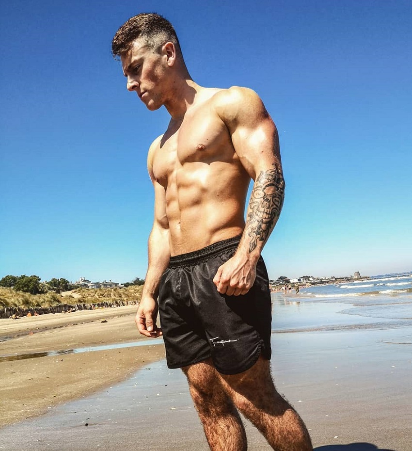 Glen Gillen posing shirtless on a beach looking muscular