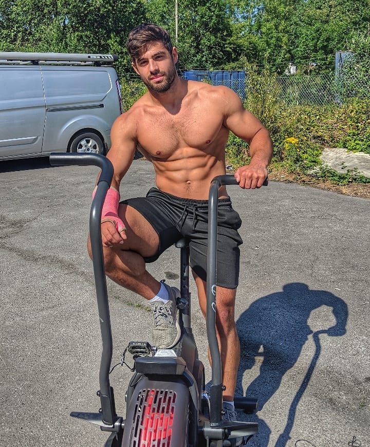 Dan Tai sitting shirtless on a cardio bike machine outdoors