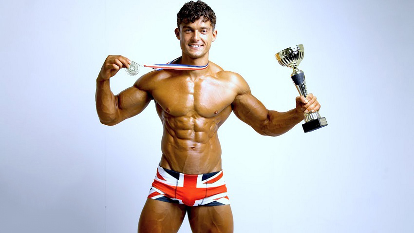 Josh Watson posing shirtless with a medal around his neck and a fitness trophy in his arm