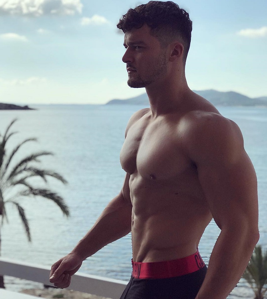 Josh Watson standing shirtless on the balcony overlooking a vast blue sea with islands