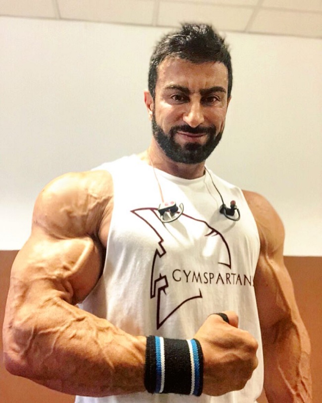 Murat Demir posing in a white tank top flexing his ripped and vascular arm