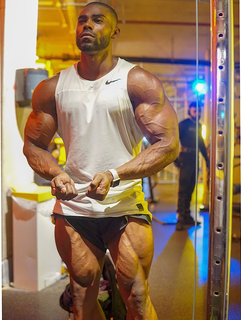 Nathan Williams flexing in a white tank top and shorts, looking huge and ripped