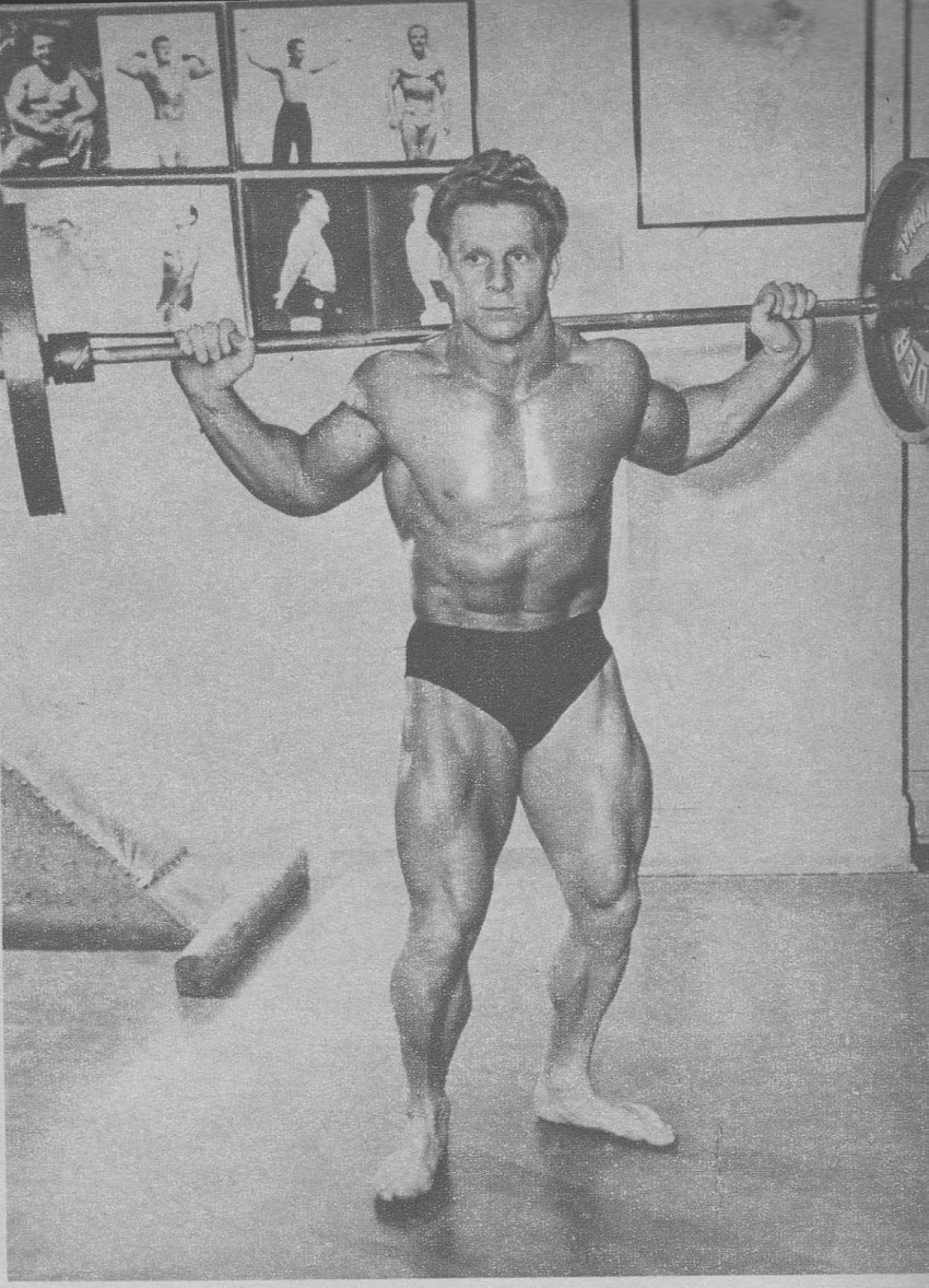 Jack Delinger standing shirtless with a heavy barbell on his back