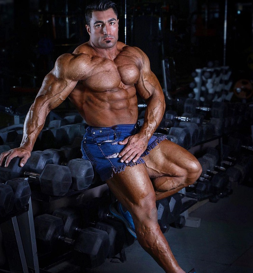 Rouhollah Mirhoseini sitting on a rack of dumbbells looking huge and swole