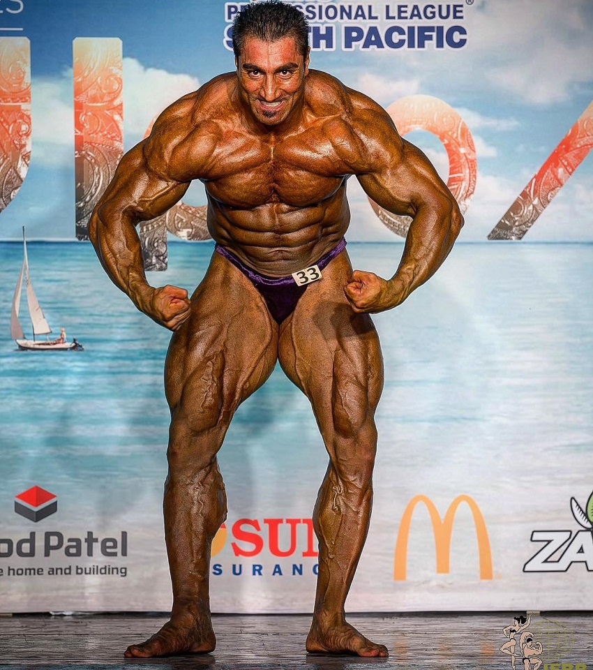 Rouhollah Mirhoseini posing on a bodybuilding stage looking huge and ripped