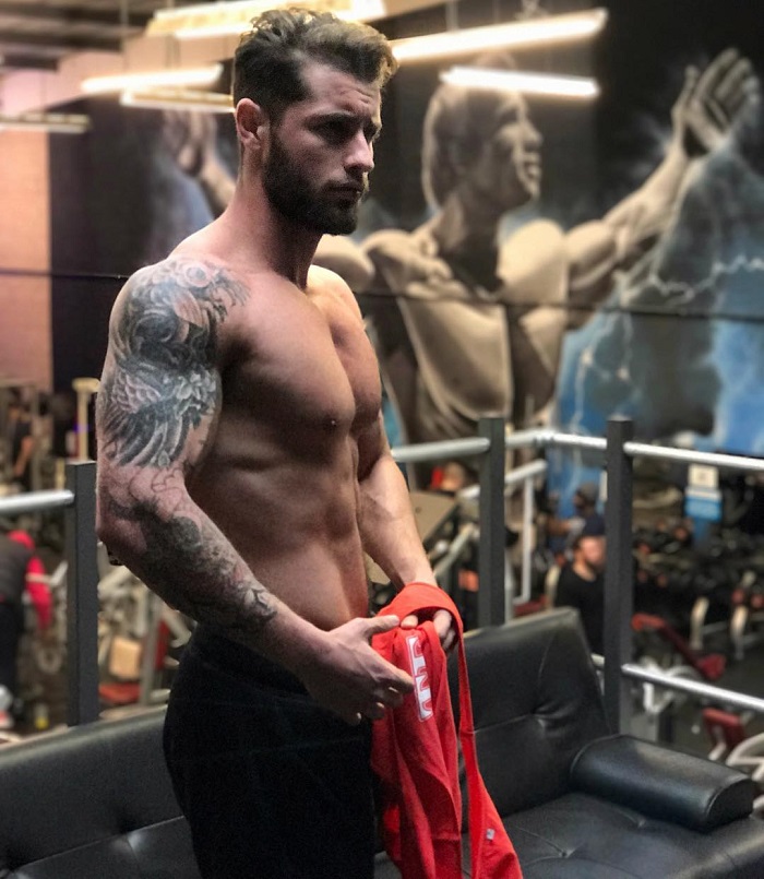 Vince Azzopardi posing shirtless in a gym with a picture of Arnold Schwarzenegger in the background