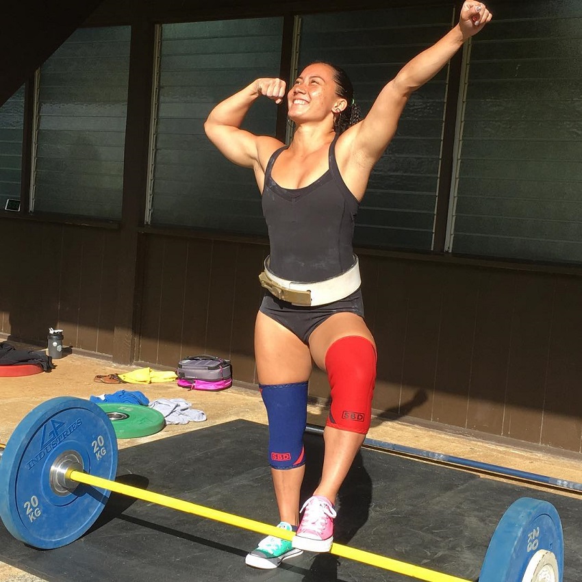 Stacia-Al Mahoe standing on a barbell with one leg and flexing her biceps