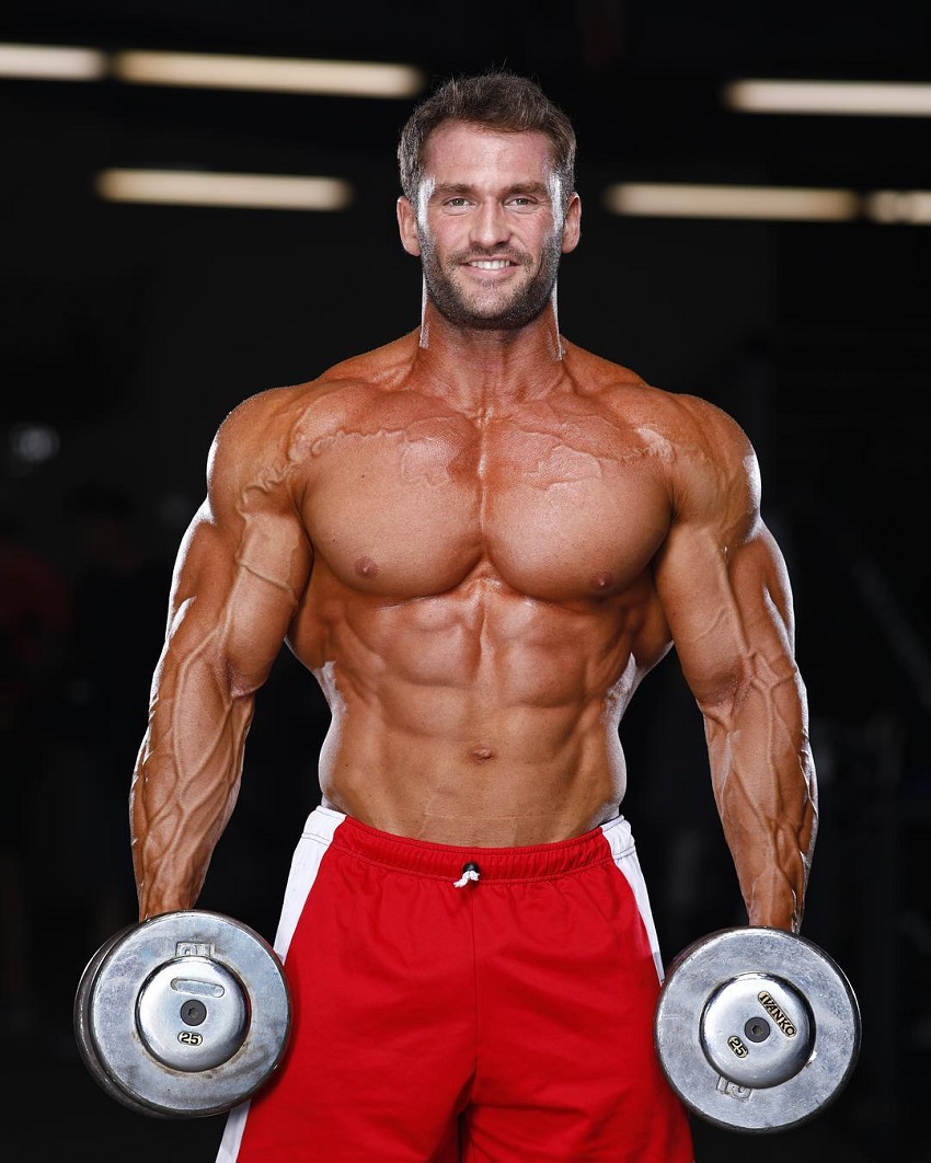 Stanimal De Longeaux standing shirtless with a dumbbell in each hand