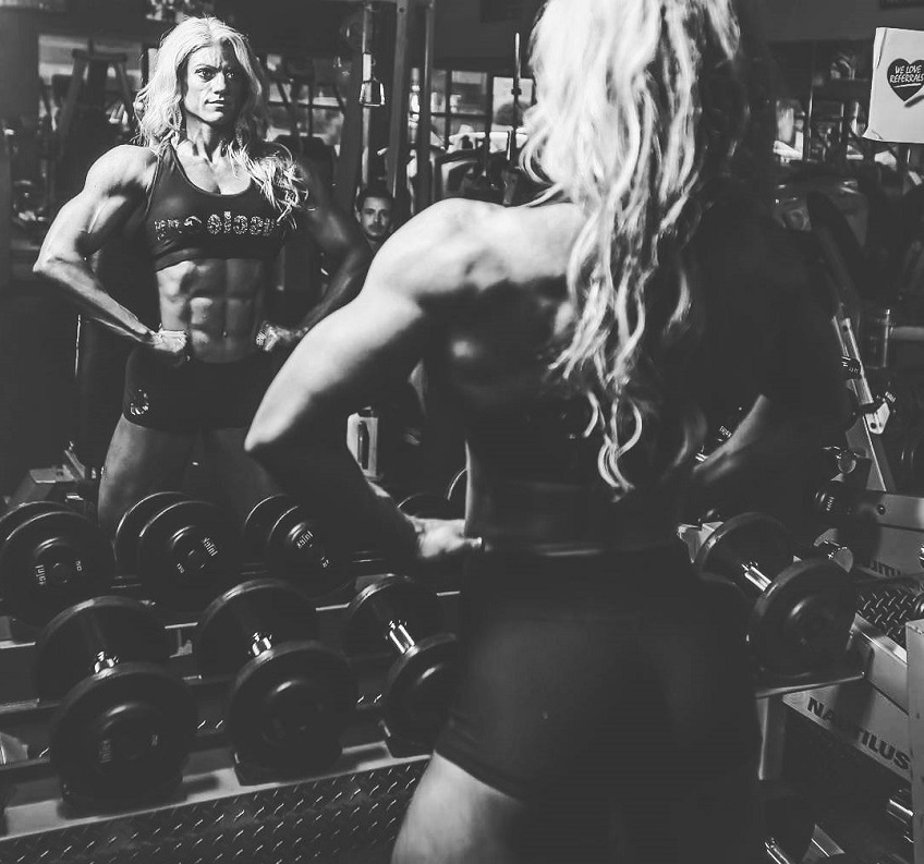 Autumn Swansen spreading her lats wide while looking at herself in front of a gym mirror