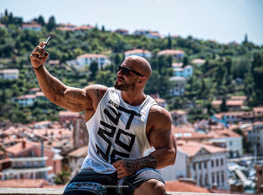 Benjamin Radic wearing a white tank top, taking a selfie of his muscular arms in an exotic location