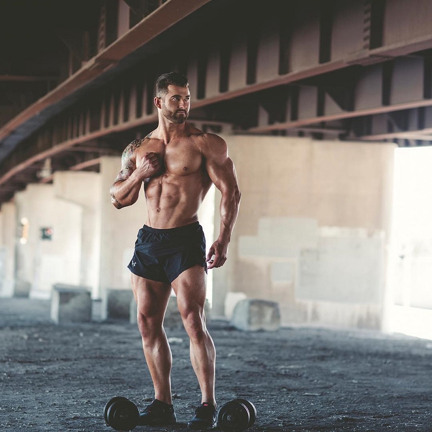 Jase Stevens posing with dumbbells by a bridge, looking ripped