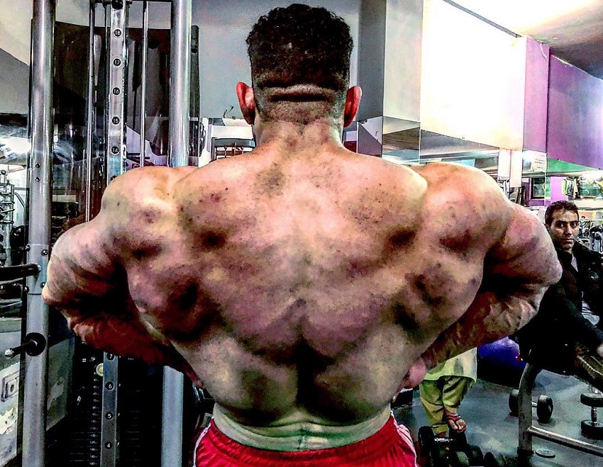 Yasin Qaderi doing a shirtless rear lat spread in a gym looking huge