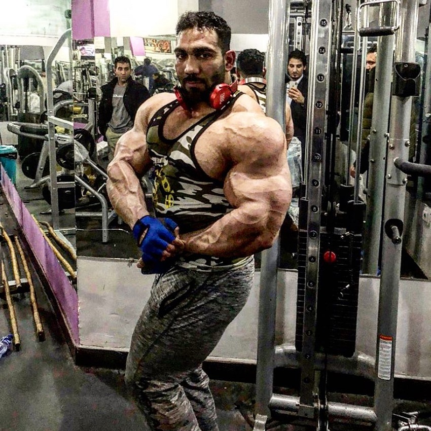 Yasin Qaderi performing a side chest pose in a gym, wearing a camouflaged tank top