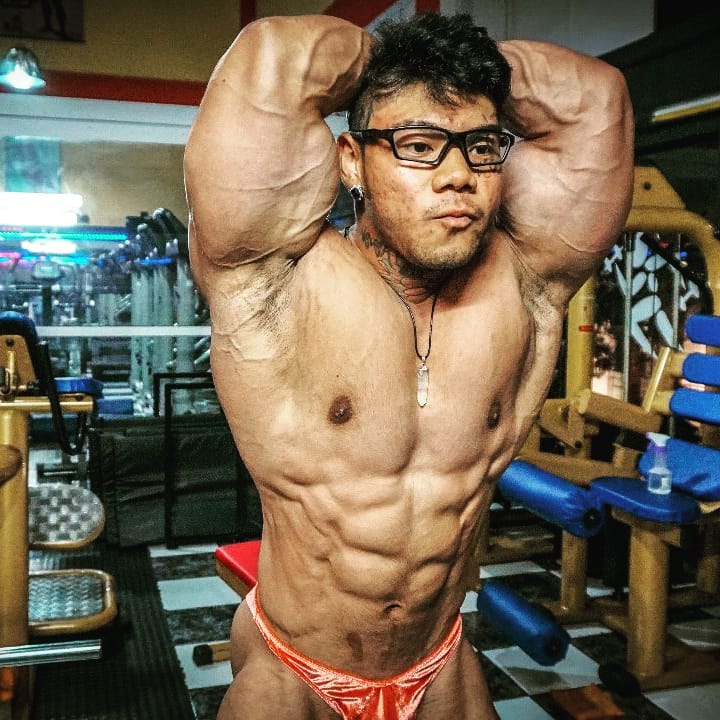 Caio Eiji Sirahata standing shirtless in the gym flexing his ripped and bulging abs