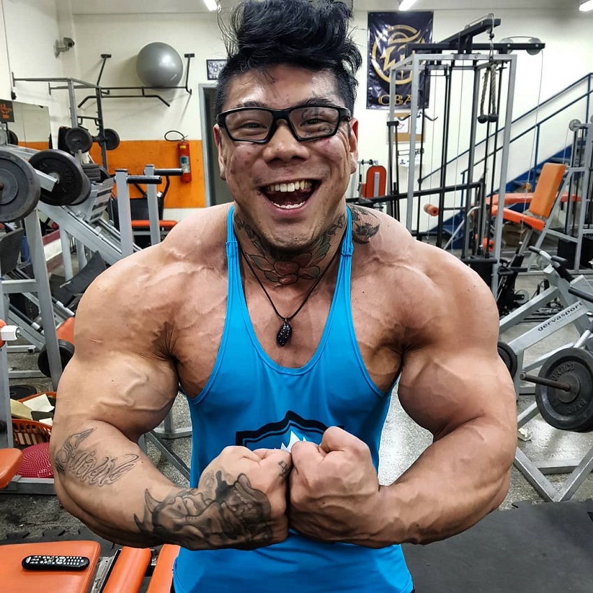 Caio Eiji Sirahata flexing for the camera, wearing a blue tank top, standing in front of cardio machines in the gym