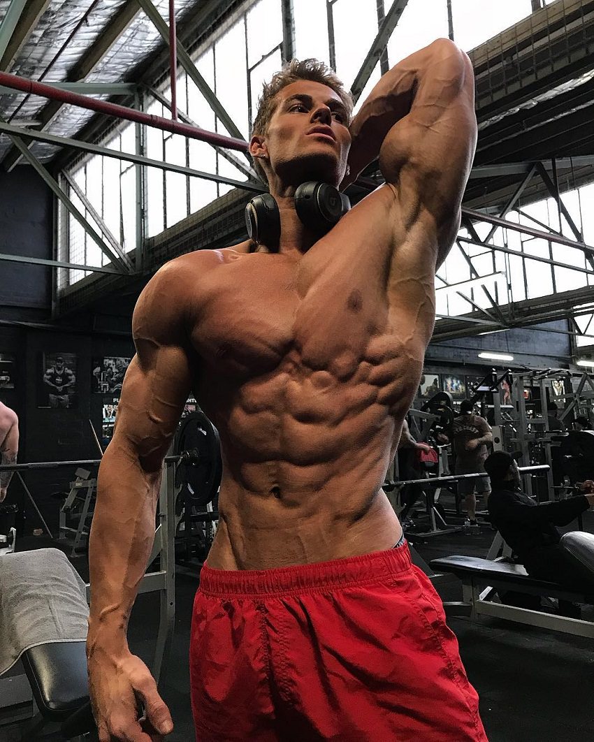 Carlton Loth posing shirtless in red shorts in the gym looking ripped and aesthetic