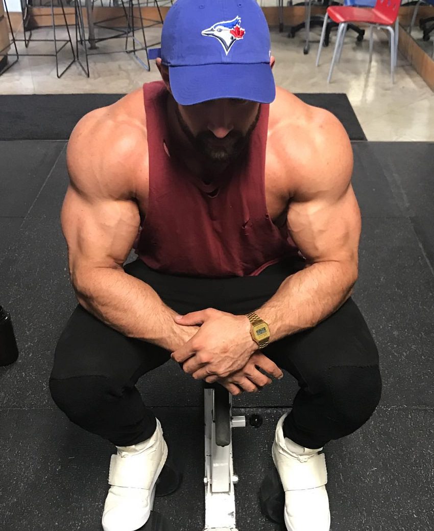 Dylan Thomas sitting on a gym bench in a red tank top, his arms looking swole and vascular