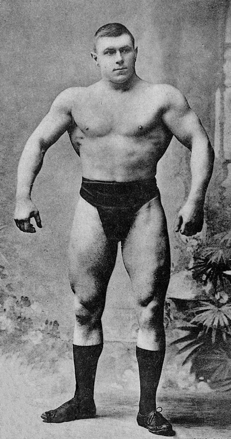 George Hackenschmidt standing shirtless, posing with his lats spread wide, looking muscular and strong