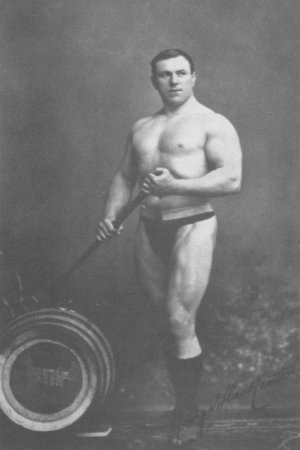 George Hackenschmidt standing shirtless next to a tyre, looking strong and muscular