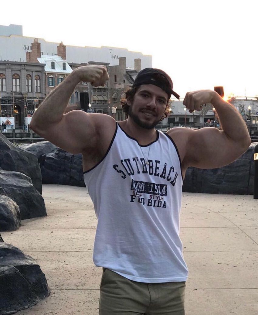 Lucas Giovani flexing hic biceps for the camera, wearing a white tank top, looking fit and muscular
