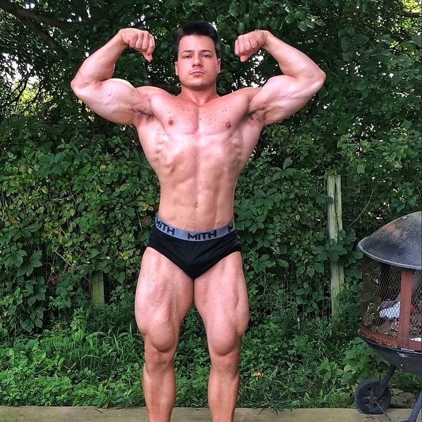 Caio Bottura performing a shirtless front double biceps pose in his backyard, looking ripped and muscular