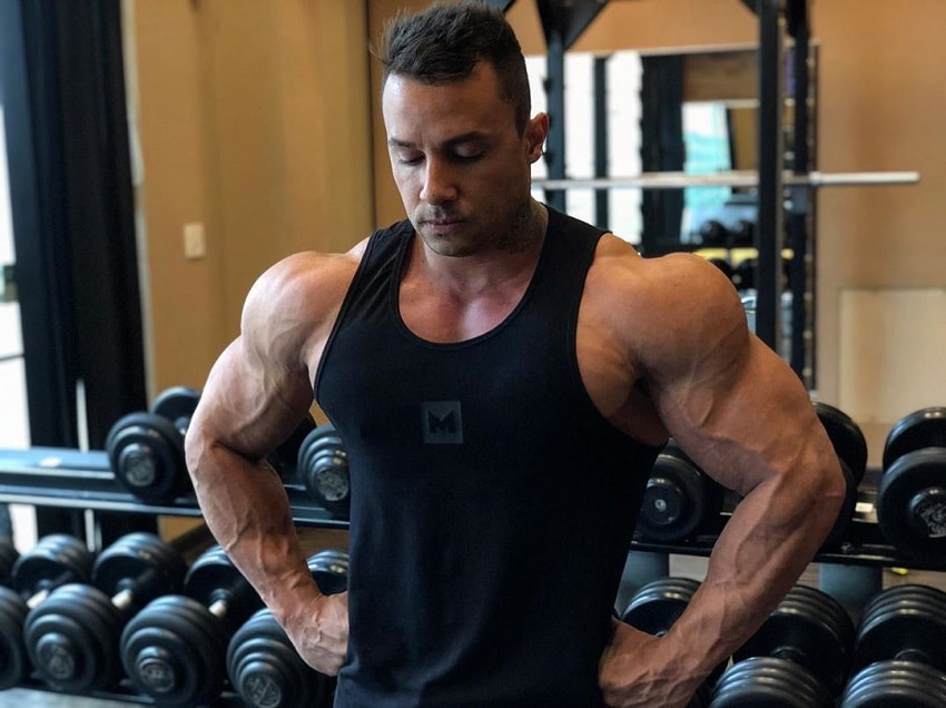 Diogo Montenegro wearing a black tank top, posing for the camera in front of the dumbbell rack, looking ripped and swole