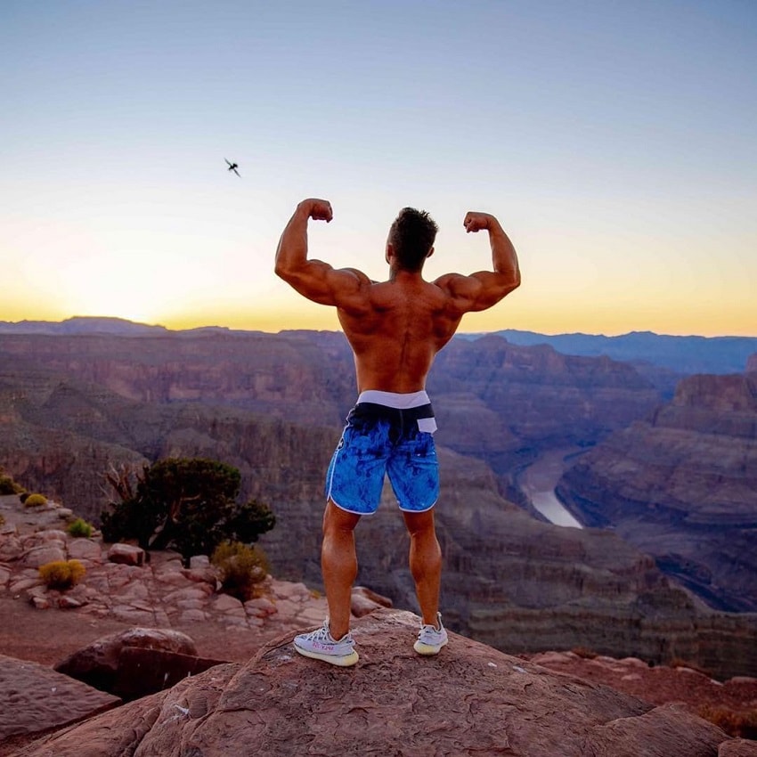 Diogo Montenegro doing a shirtless back double biceps outdoors on a barren hill overlooking a huge canyon