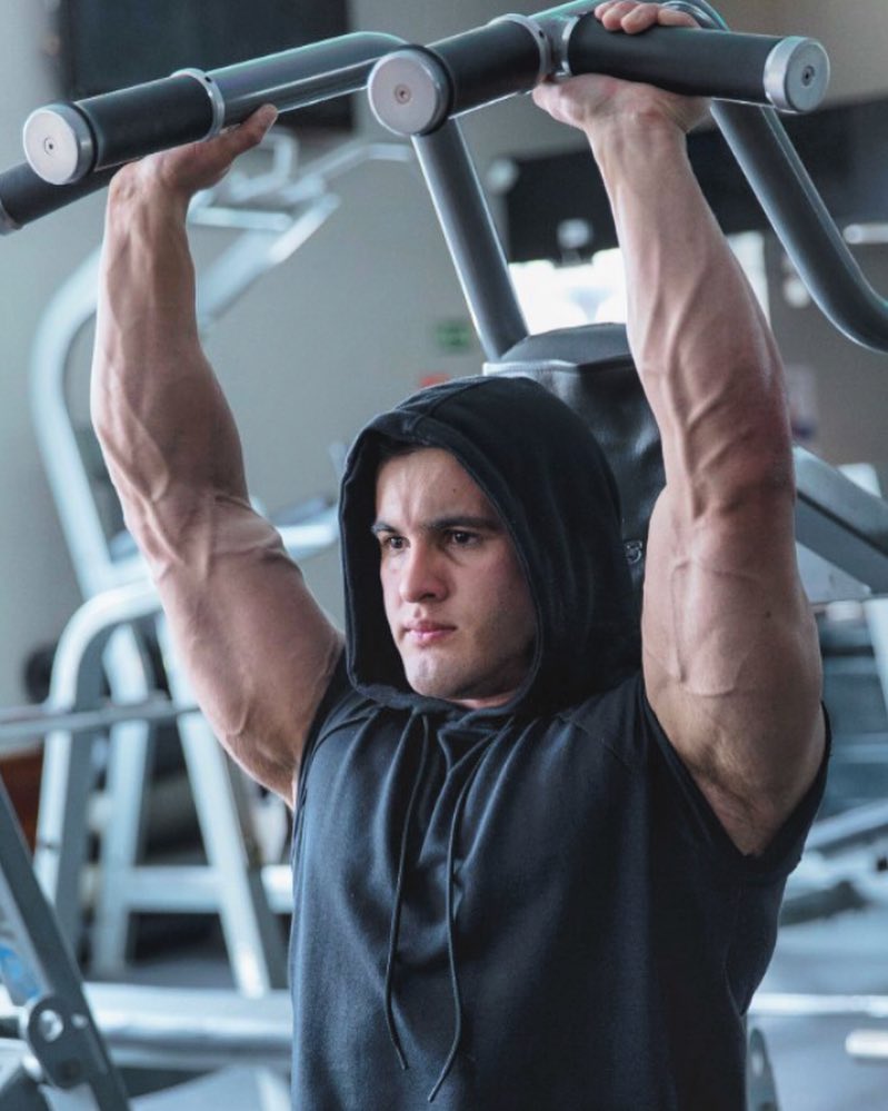 Ernane Guimaraes lifting heavy overhead press on the machine in the gym, looking vascular and ripped