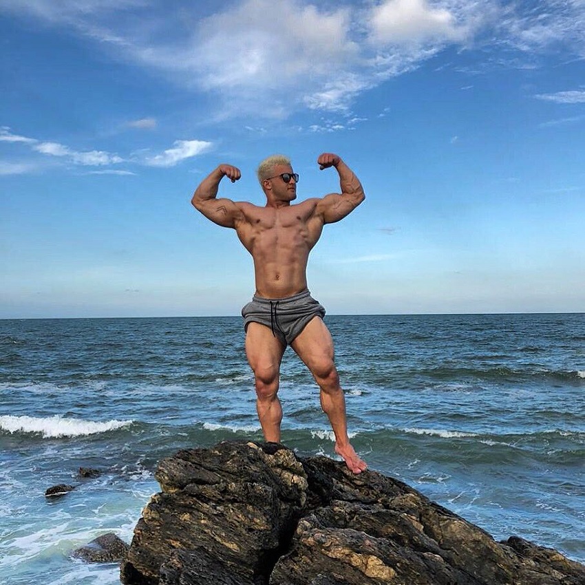 Ernane Guimaraes flexing front double biceps on the rock by the sea