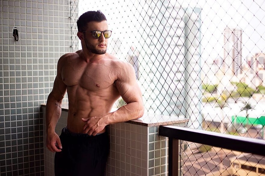 Ernane Guimaraes posing shirtless on the balcony with sunglasses on, looking fit and ripped