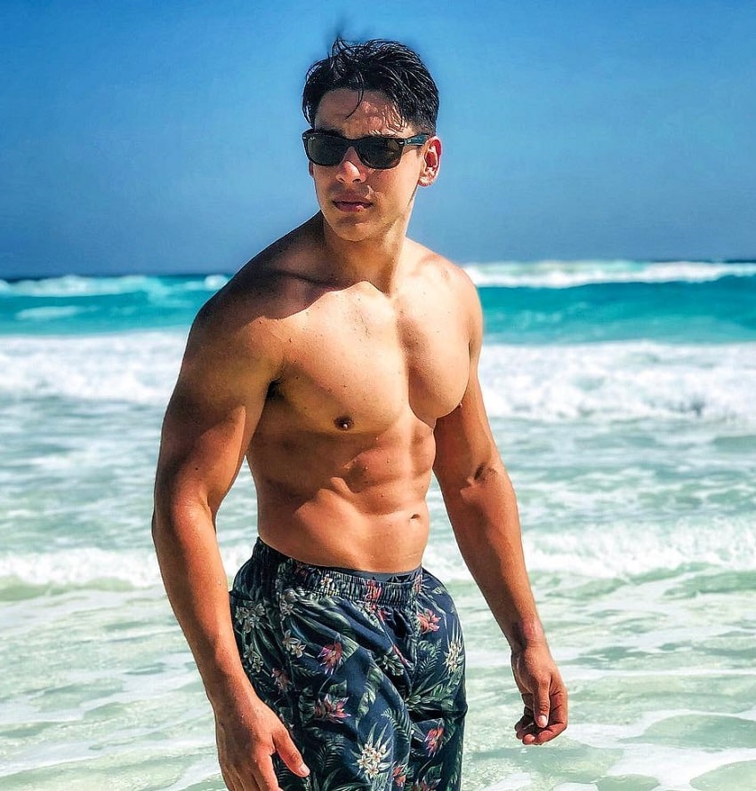 Gabriel Arones standing shirtless on the beach with black sunglasses, looking fit and strong