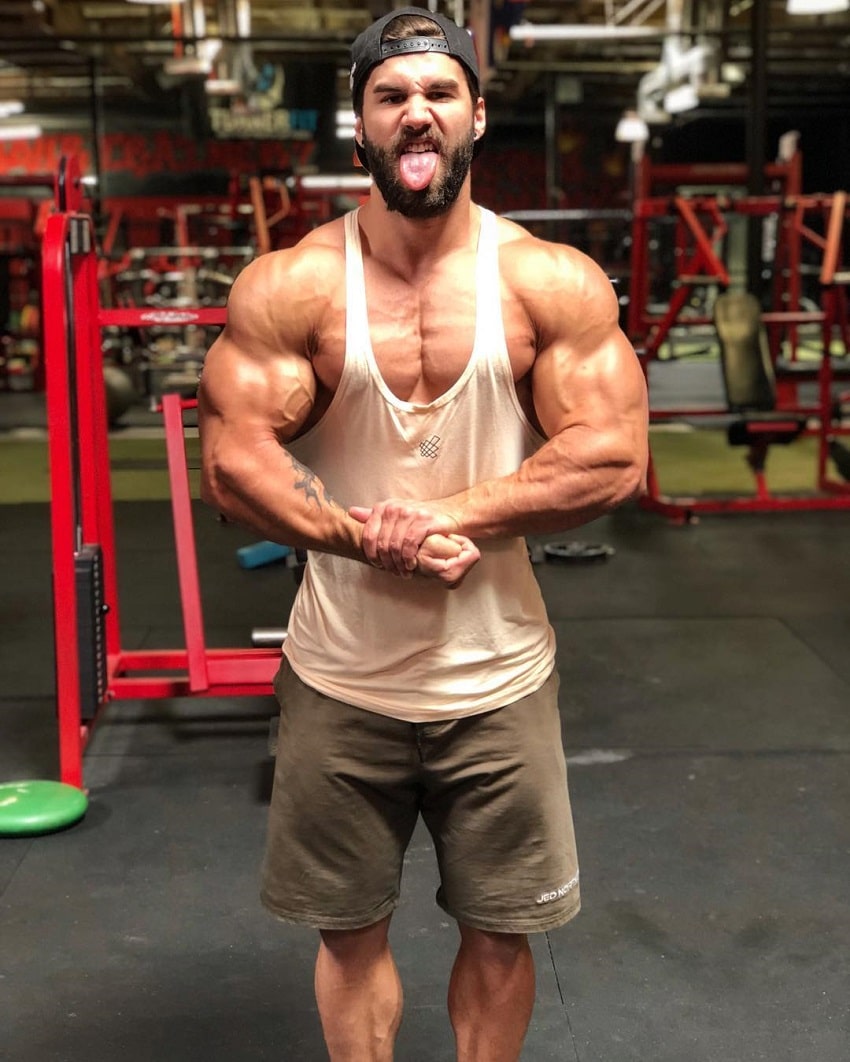 Jake Burton performing a most muscular pose in his white tank top while looking at the camera, looking strong and ripped