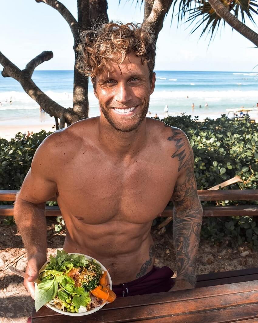 Matt Fox sitting shirtless in the shade near a beach, showing off his plate with vegan food